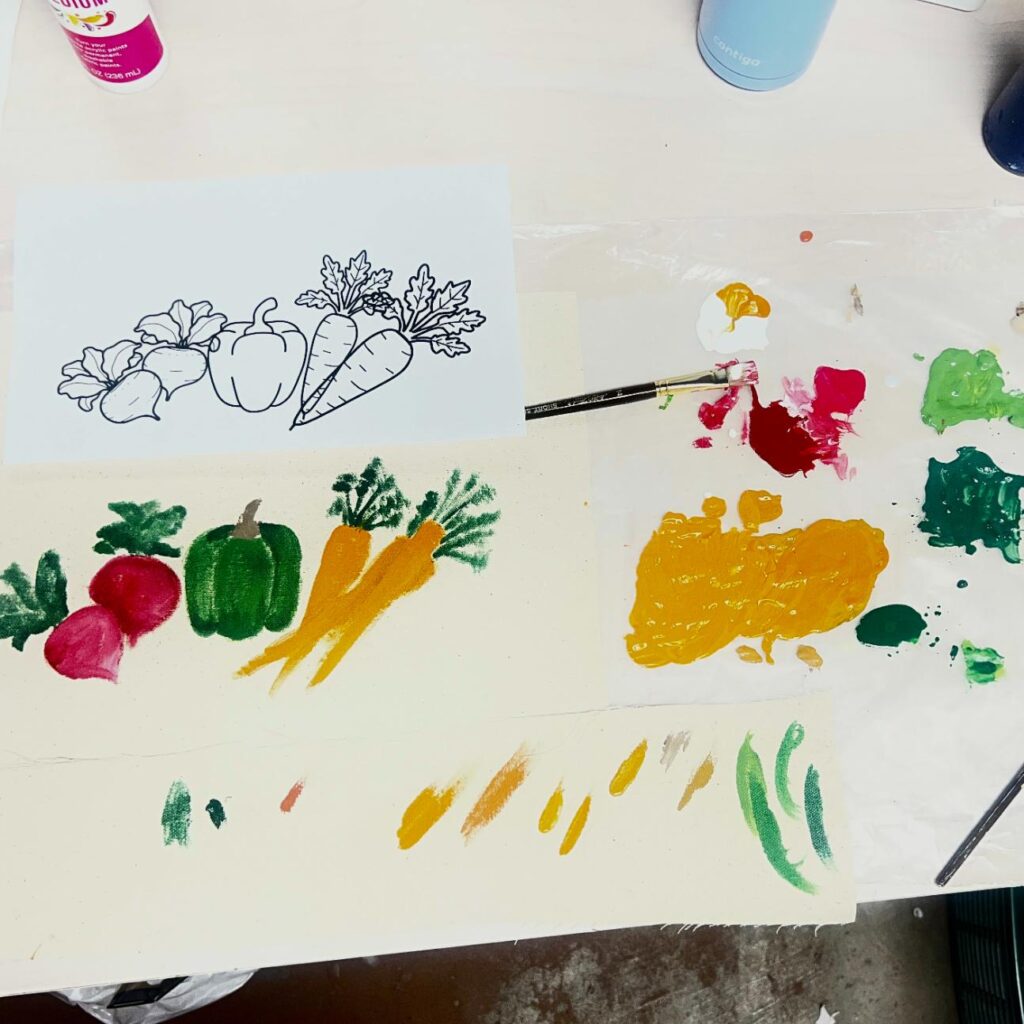 A workspace with a vegetable coloring page, paint palette, brush, and painted radishes, bell pepper, and carrots on a canvas.