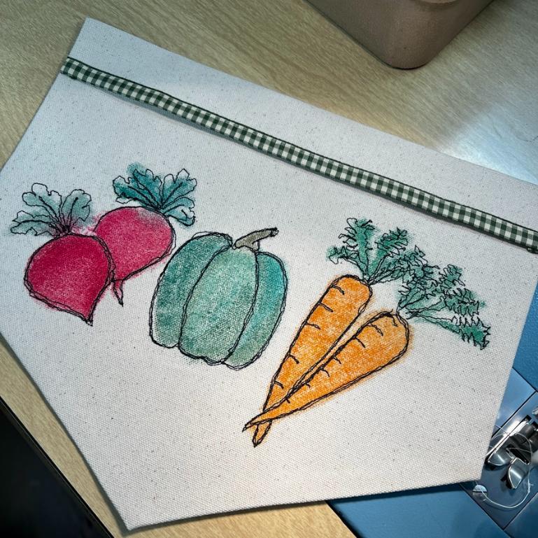A fabric pennant featuring embroidered and colored images of two beets, a green squash, and two carrots, with a green checkered ribbon sewn at the top, lying on a wooden surface.