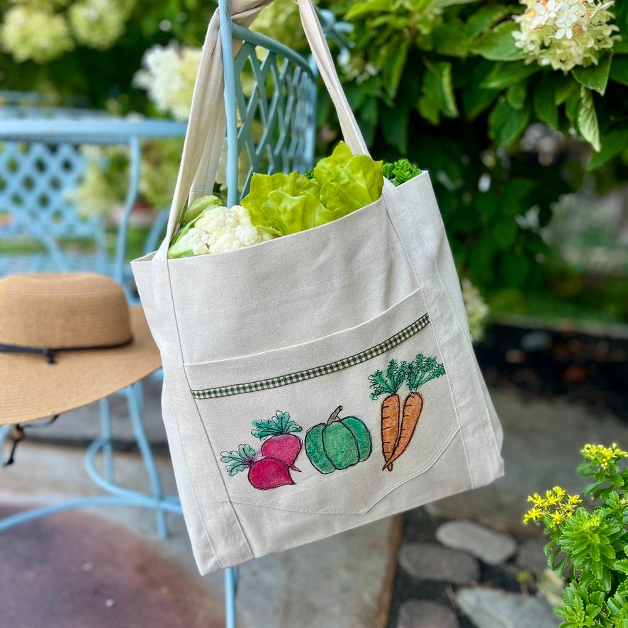 A white tote bag with embroidered vegetables holds fresh lettuce and cauliflower. The bag is on a blue chair outdoors near a straw hat and green plants.