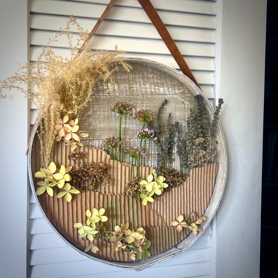 A circular wall hanging with dried flowers and grasses, arranged on corrugated cardboard, is suspended by a brown leather strap against a white louvered background—a charming wreath alternative for adding rustic elegance to your space.