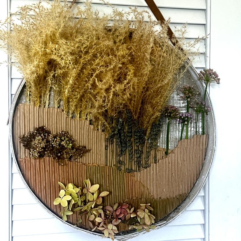 A round wall art piece, serving as a wreath alternative, features dried grasses, flowers, and leaves arranged on textured cardboard to resemble a natural landscape, hanging against white shutters.