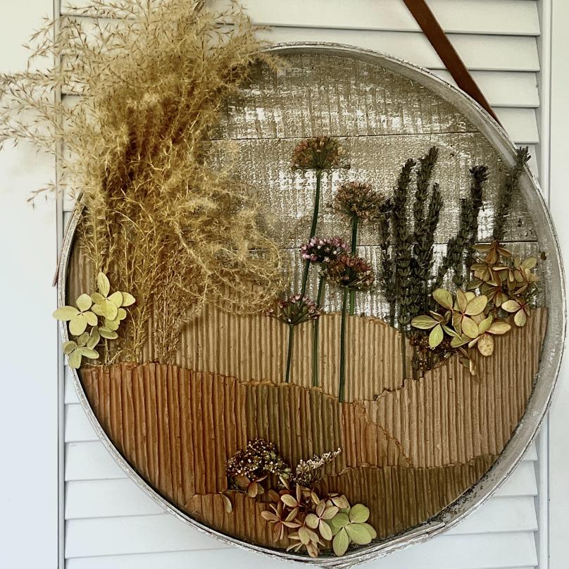 A round wall art piece serves as a stunning wreath alternative, featuring dried grasses, flowers, and leaves arranged on corrugated cardboard. This creates a textured landscape scene with earthy tones, beautifully mounted against a white slatted background.
