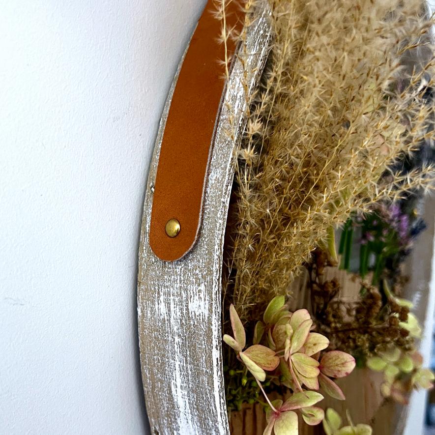 A close-up of a rustic, whitewashed wooden wall decor piece with a brown leather strap and dried flowers, including grasses and hydrangeas, arranged inside—offering a stylish wreath alternative for your home.