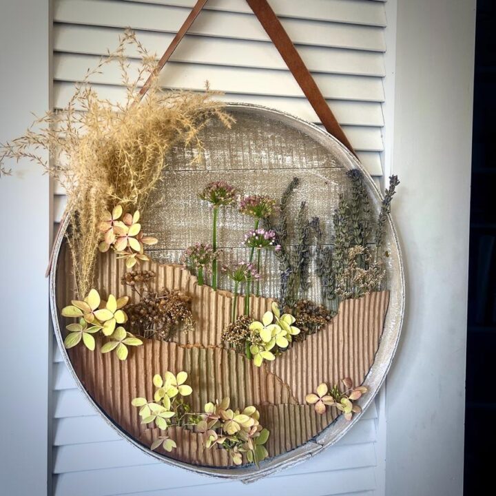 A circular wall hanging with dried flowers and grasses arranged on a textured background, featuring earthy colors and a brown leather strap for hanging—perfect as a stylish wreath alternative.