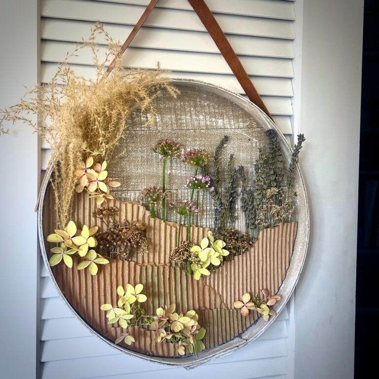 A circular wall hanging with dried flowers and grasses arranged on a textured background, featuring earthy colors and a brown leather strap for hanging—perfect as a stylish wreath alternative.