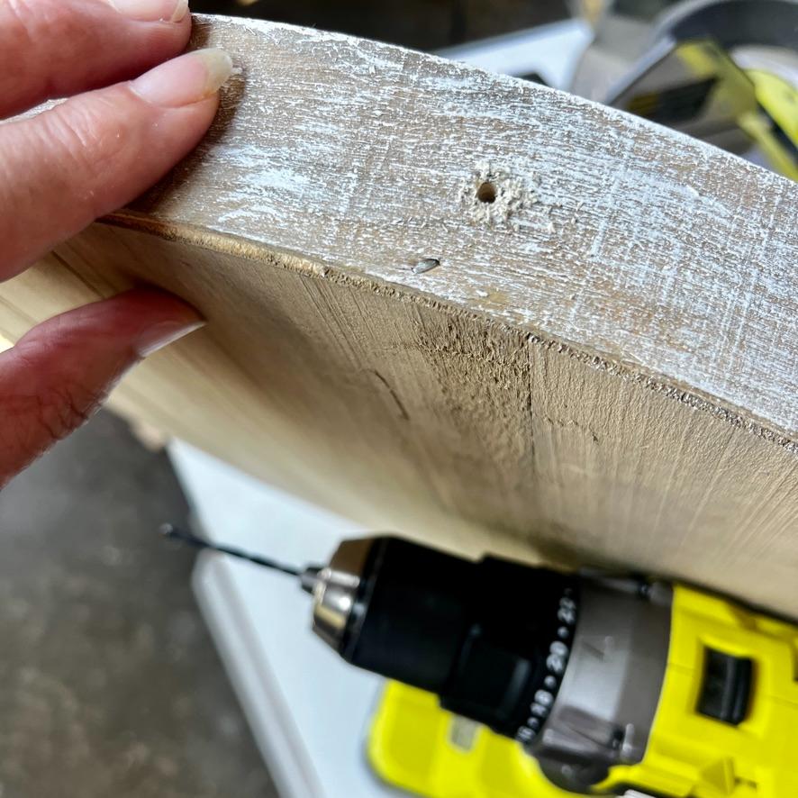 A hand holds a wooden board with a drilled hole near the edge—an ideal base for a creative wreath alternative. A yellow and black power drill with a bit attached rests below the board.