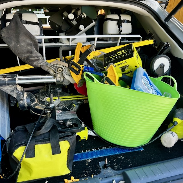 An open car trunk filled with construction tools, including a saw, a green tub, a yellow and black tool bag, levels, and various cases and equipment. The back seats are visible in the background.