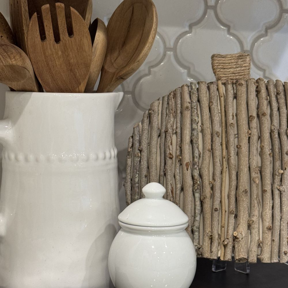 A white ceramic pitcher holds wooden utensils next to a round trivet made of vertical wooden sticks and a small white lidded jar, all set against a white tile backsplash—a charming scene that offers a natural wreath alternative for your kitchen decor.