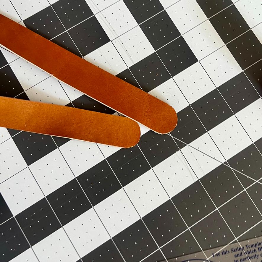 Two brown leather straps with rounded ends—perfect as a wreath alternative—are placed diagonally on a black and white grid cutting mat with measurement markings.