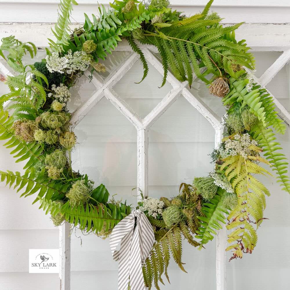 A lovely wreath alternative made of green ferns and small flowers hangs on a white glass-paneled door. A striped ribbon bow is tied at the bottom of the wreath, creating a fresh and unique look. The background is light and airy.