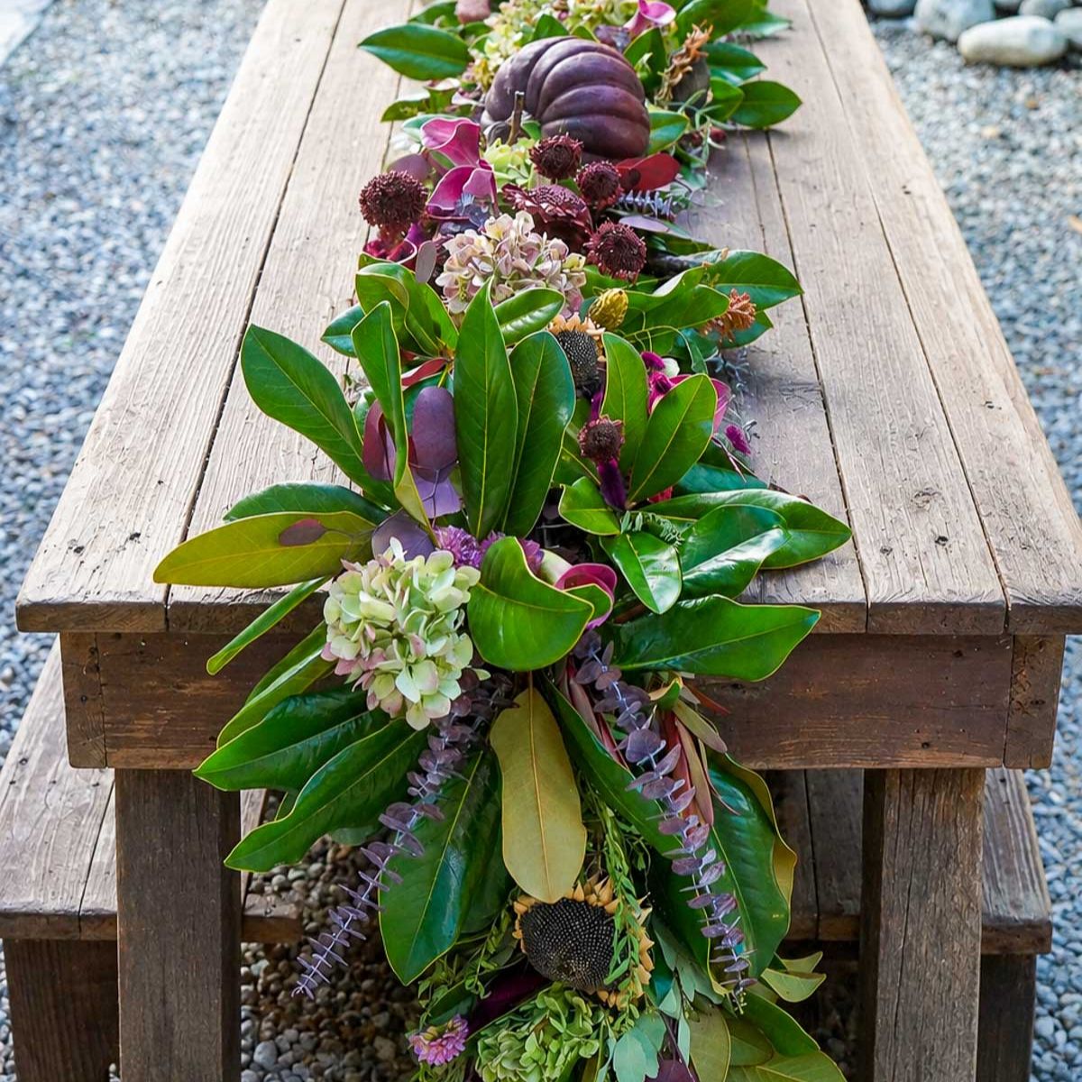 A rustic wooden table is decorated with a fall garland of greenery, purple leaves, hydrangeas, dried flowers, and a small purple pumpkin—serving as a stunning wreath alternative and creating an autumn centerpiece. Pebbles surround the outdoor setting.