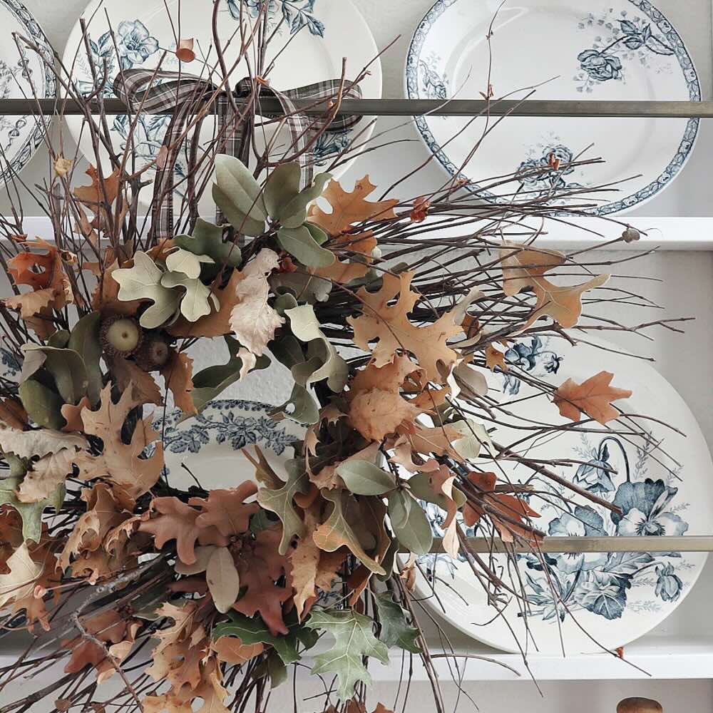 A wreath alternative made of dried leaves and twigs hangs in front of a set of white plates with blue floral designs displayed on a shelf.