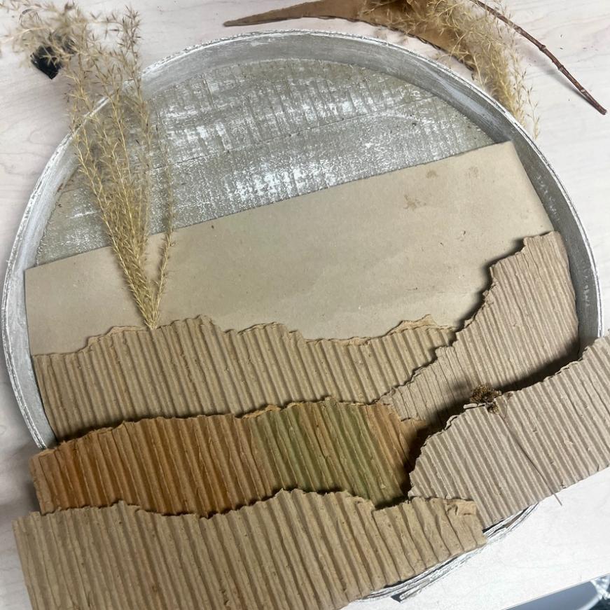 A circular tray with corrugated cardboard pieces arranged like mountains, dried grasses, and a sheet of paper creates a textured landscape art scene—an inventive wreath alternative for showcasing natural beauty in any space.