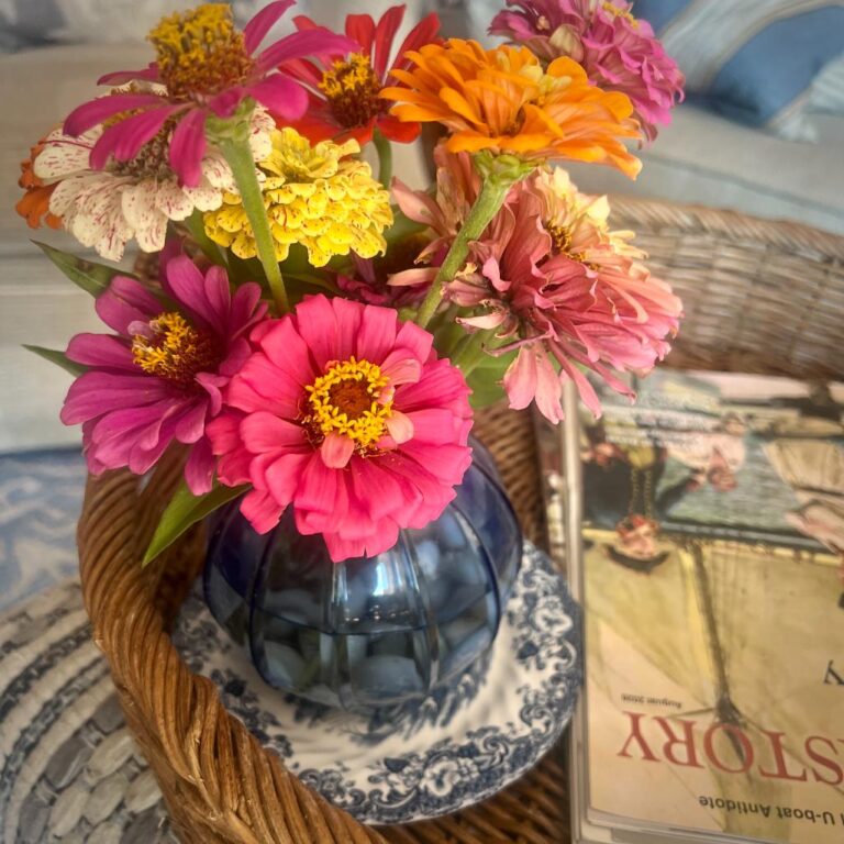 A small blue vase with colorful zinnias sits on a decorative plate inside a wicker basket, next to a magazine titled “STORY” on a table.