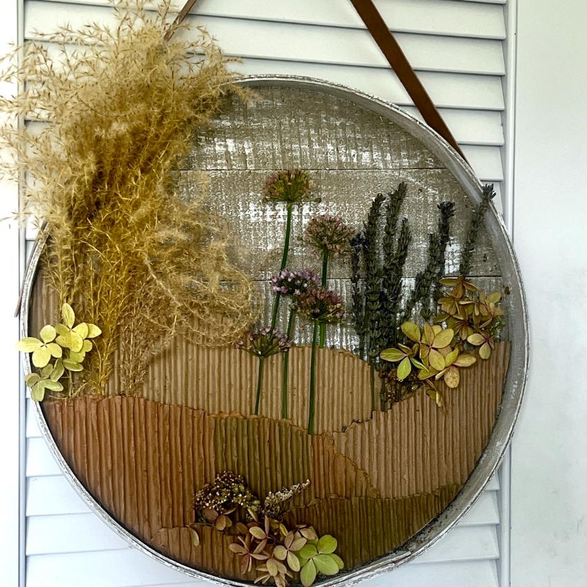 A round wall art piece with dried grasses, flowers, and leaves arranged within a shallow metal tray, hanging on a white shuttered background. Offering a unique wreath alternative, the design resembles a natural landscape scene.