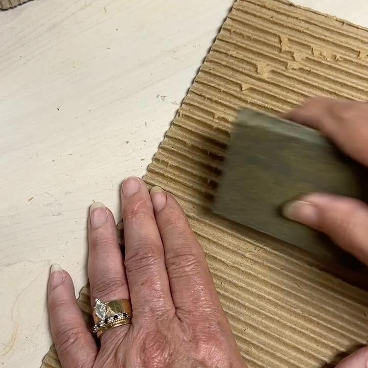 A person uses a sanding block on a piece of corrugated cardboard, possibly crafting a wreath alternative, placed on a light-colored surface. One hand holds the cardboard steady, showing rings on their fingers.