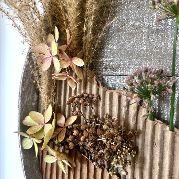 A close-up of dried flowers, grasses, and green stems arranged on a rustic wooden surface with a piece of corrugated cardboard—an artful wreath alternative for natural decor lovers.