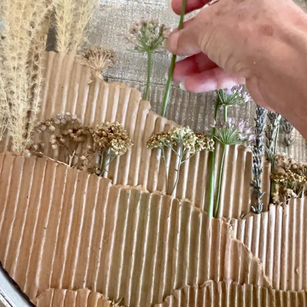 A hand arranges dried flowers and grasses on a textured, corrugated cardboard background, creating a layered, nature-inspired art piece that serves as a unique wreath alternative.