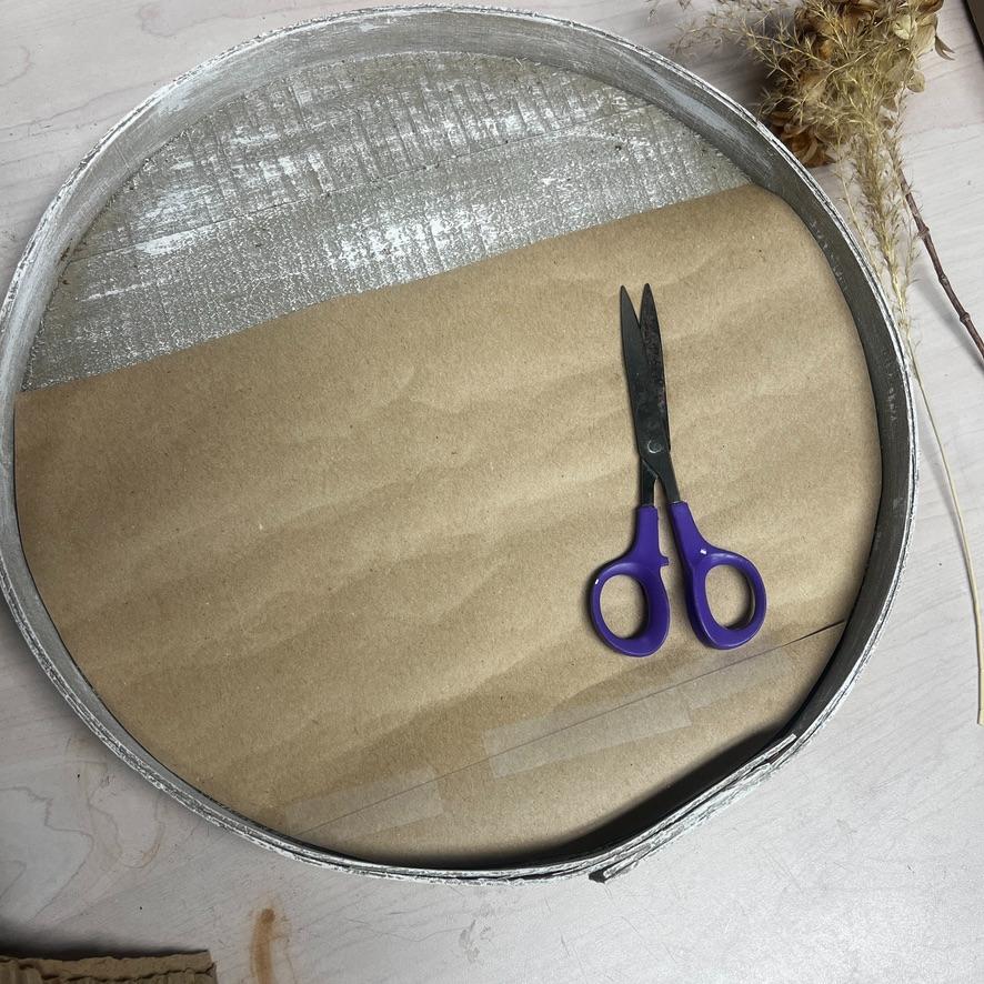A metal ring with brown paper inside, purple-handled scissors on top, and dried plants nearby creates a charming wreath alternative, all arranged on a light-colored table.
