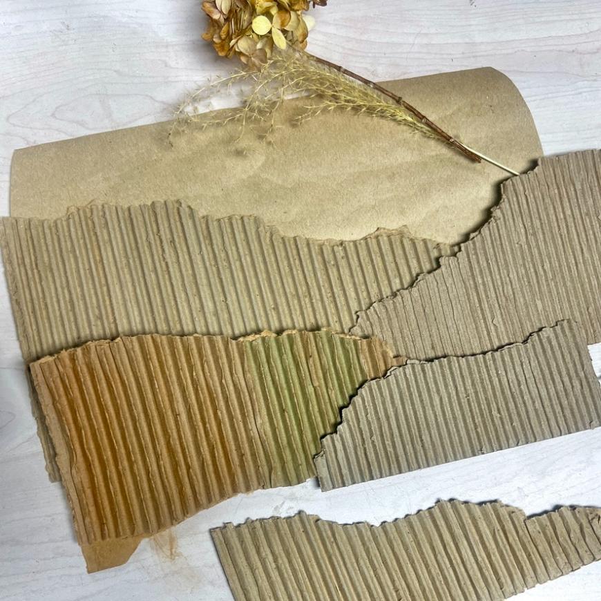 Torn pieces of corrugated cardboard and brown kraft paper are arranged on a white surface, with dried flowers placed at the top, creating a rustic wreath alternative for your decor.