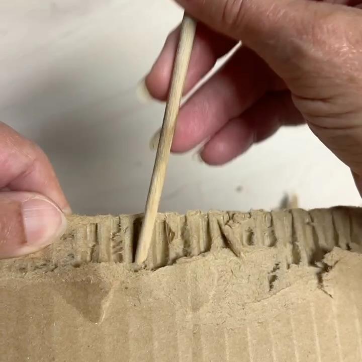A hand uses a wooden stick to separate the layers of corrugated cardboard, revealing the wavy inner structure—an inventive approach that could inspire a unique wreath alternative.