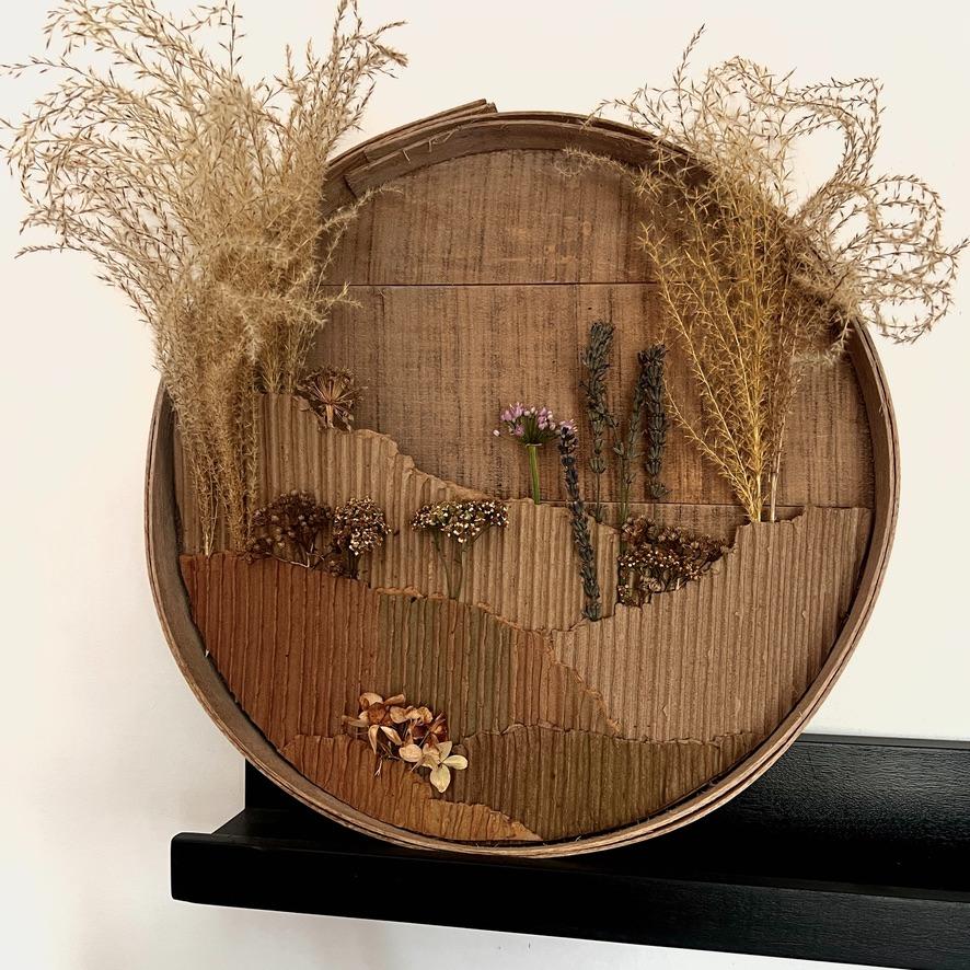 A round, textured cardboard art piece with dried grasses and flowers arranged to resemble a natural landscape serves as a unique wreath alternative, displayed on a black shelf against a white wall.