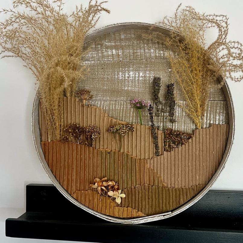 Round wall art made from corrugated cardboard and dried plants, depicting a textured landscape with hills, grasses, and small flowers in earthy tones, displayed on a black shelf—a unique wreath alternative that brings natural beauty to your home décor.