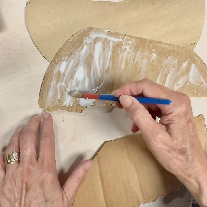 A person uses a small paintbrush to spread white glue onto a curved piece of cardboard, working on what appears to be a creative wreath alternative, with more cardboard pieces and a tabletop visible in the background.