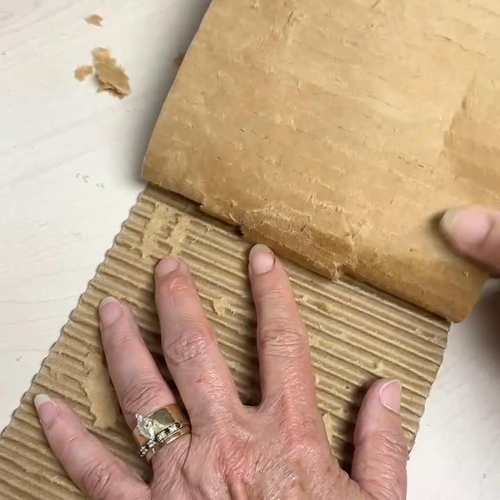 A hand peels the flat top layer off a piece of corrugated cardboard, revealing its wavy inner structure—a creative twist that could be used as a wreath alternative. The person wears two rings on their left ring finger.