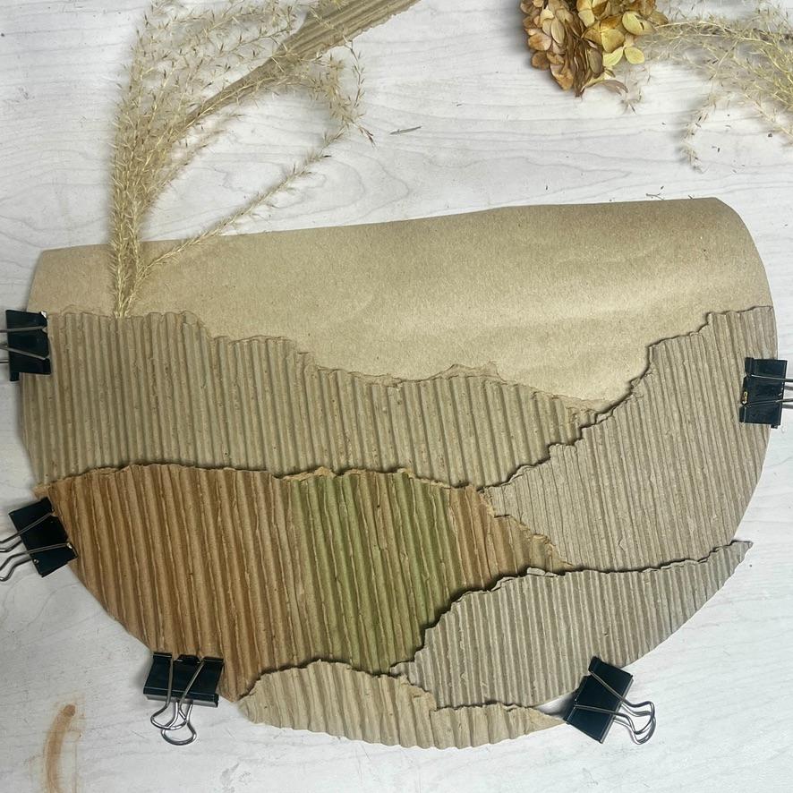 Curved pieces of corrugated cardboard layered and clipped together with binder clips, resembling a landscape, with decorative dried grasses and flowers on a light surface—a creative wreath alternative that adds natural charm to your décor.