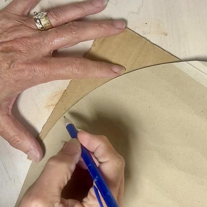 A person traces a curved line on brown paper with a blue pen, holding the paper steady with their other hand. Creating a unique wreath alternative, the person wears a ring with a stone on their left hand.