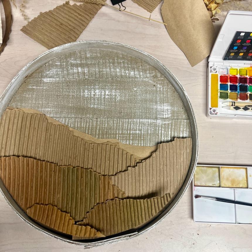 A round tin holds layered, textured cardboard pieces resembling mountains—a unique wreath alternative; nearby is a watercolor paint set and brush on a light wooden table.