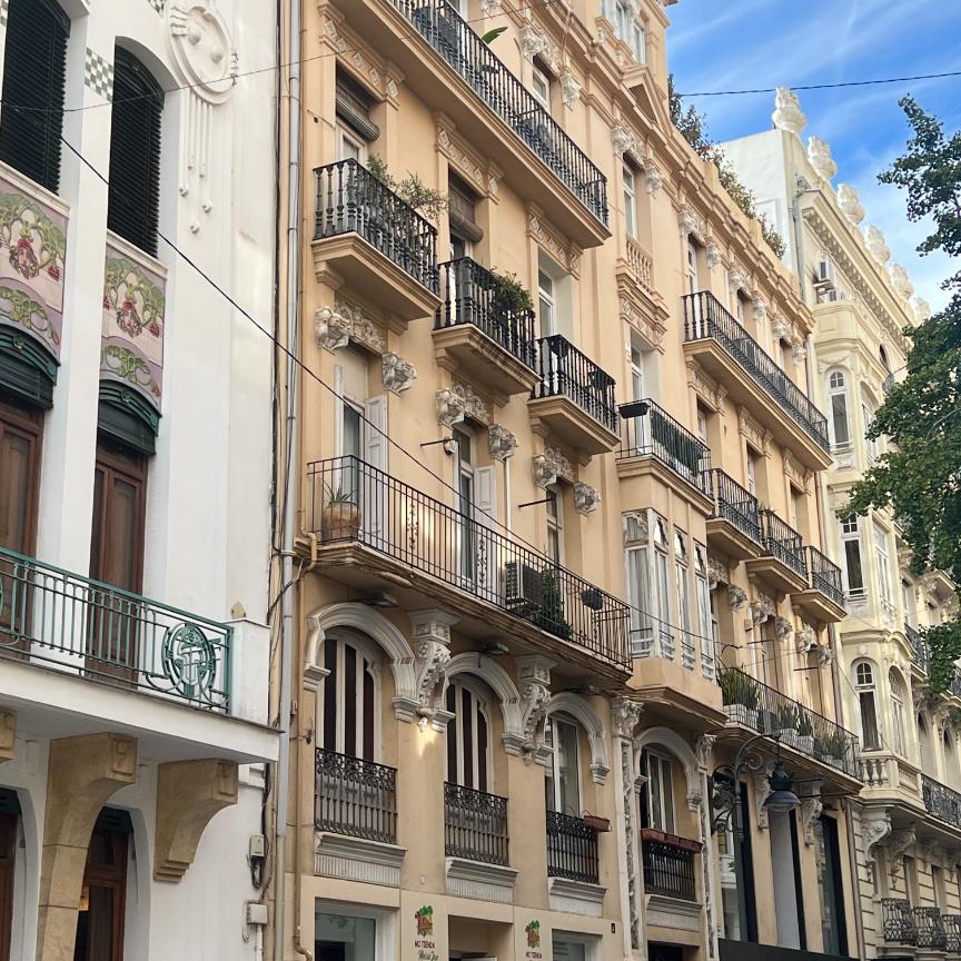 A row of ornate, multi-story buildings in a European city, featuring cream and pale yellow facades, decorative carvings, and black iron balconies.