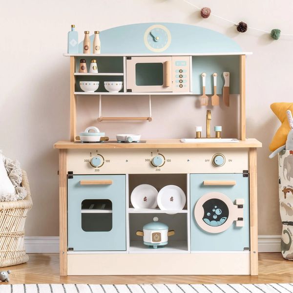 A pastel-colored wooden toy kitchen set with a stove, oven, microwave, sink, and utensils, decorated with small play food and cookware, stands against a beige wall.