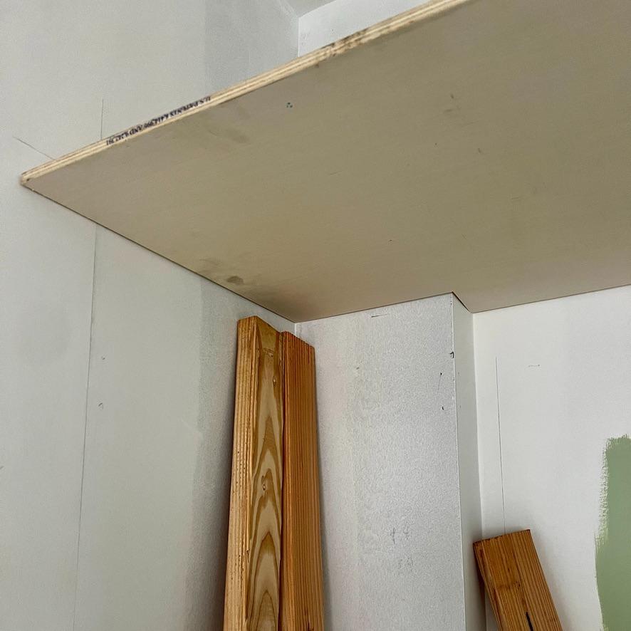A plywood shelf installed in the corner of an IKEA Pax closet wall, with two pieces of wooden trim leaning against the white wall below.