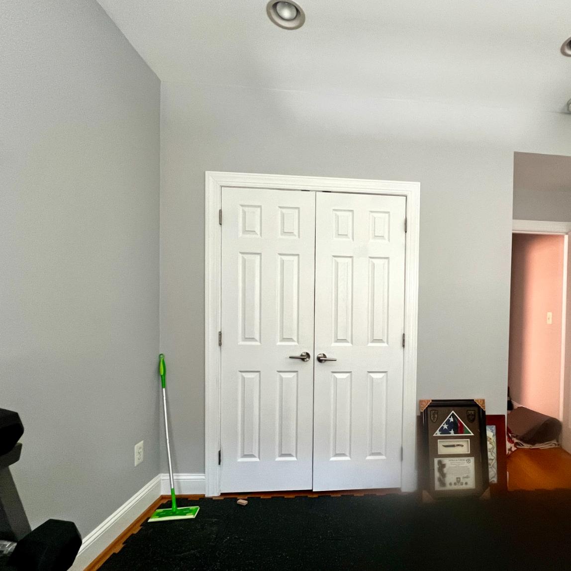 White double doors in a gray room with a floor mop standing in the corner and a framed American flag display on the floor.