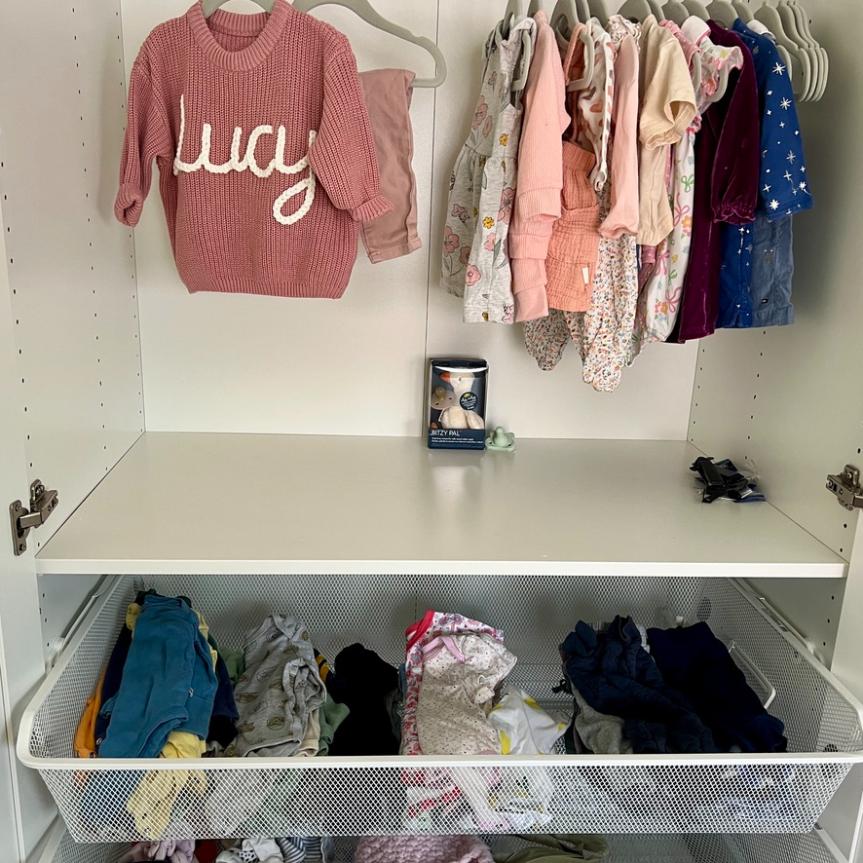 An IKEA Pax closet with baby clothes hanging on a rack above and folded baby clothes in a wire mesh drawer below.