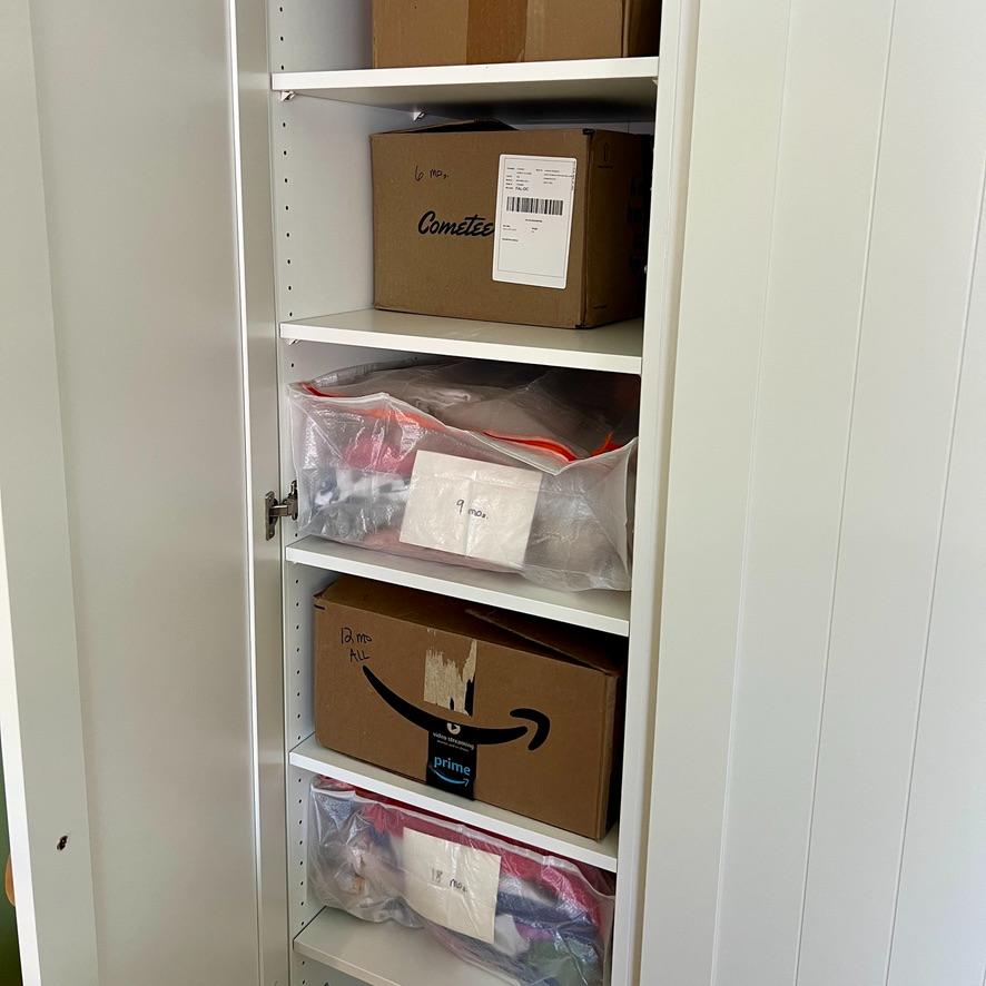 A tall white IKEA Pax closet wall holds shelves with storage boxes and clear plastic bags organized by infant clothing sizes ranging from 6 months to 18 months.