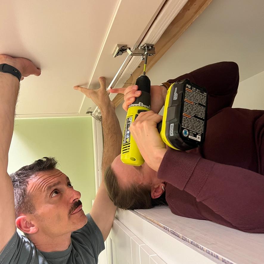 One person holds a cabinet door in place for DIY storage above Pax wardrobes while another uses a power drill to install the hinge.