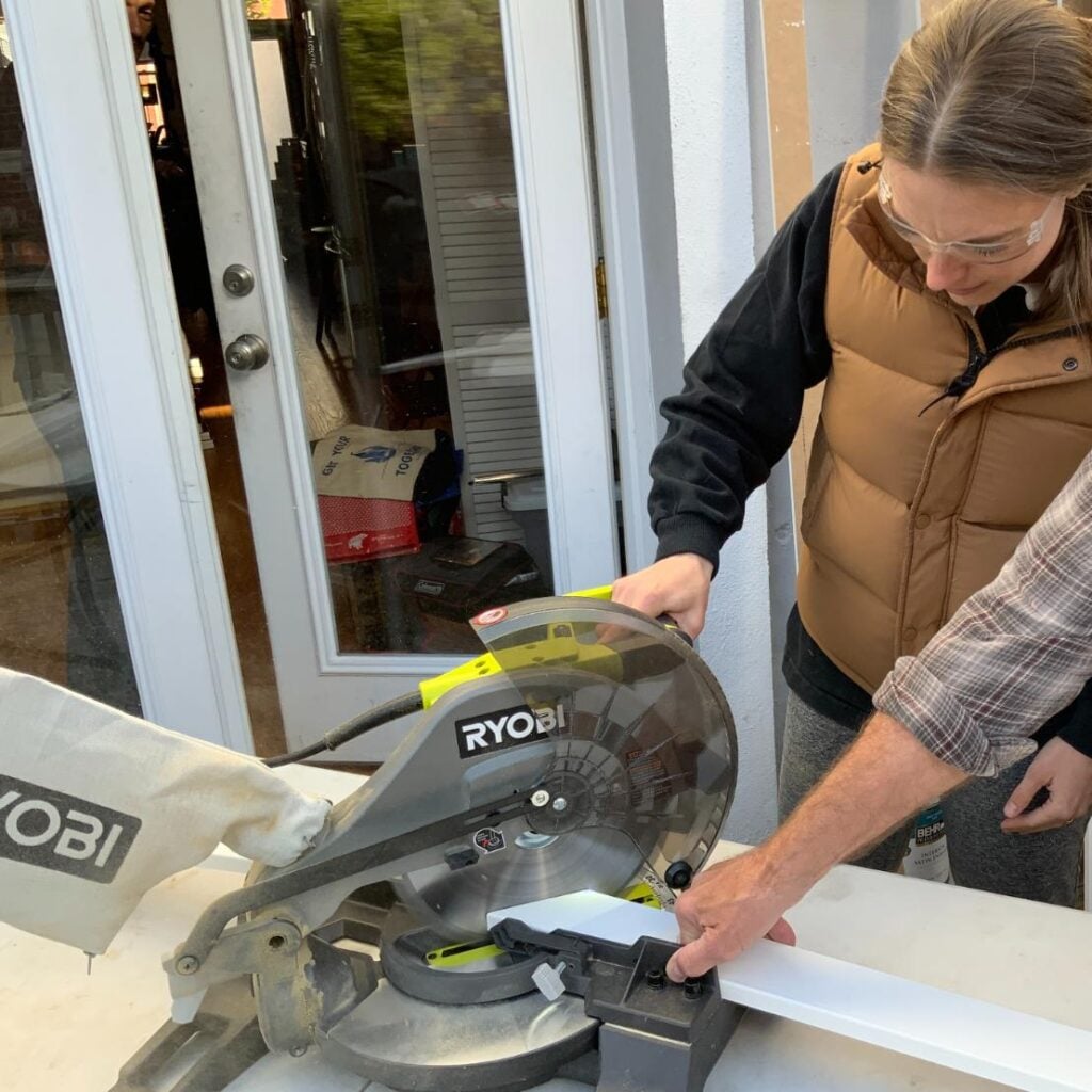 A person in a brown vest uses a Ryobi miter saw to cut white trim for DIY storage above Pax wardrobes.