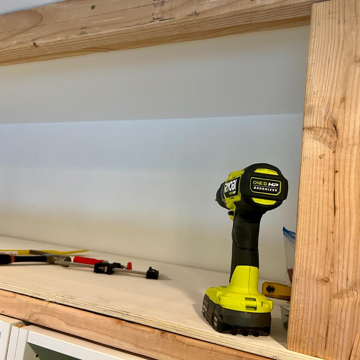 A cordless Ryobi drill stands on a plywood shelf inside a wooden frame for DIY storage above pax wardrobes, with a clamp resting nearby.