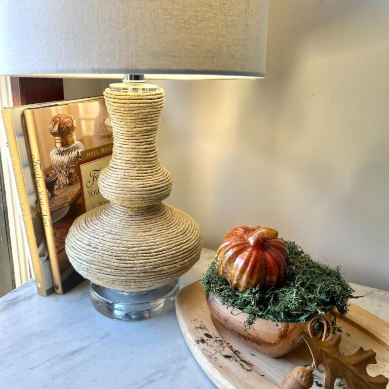 A rope-wrapped lamp stands next to two books and a small decorative pumpkin resting in a wooden bowl filled with moss.