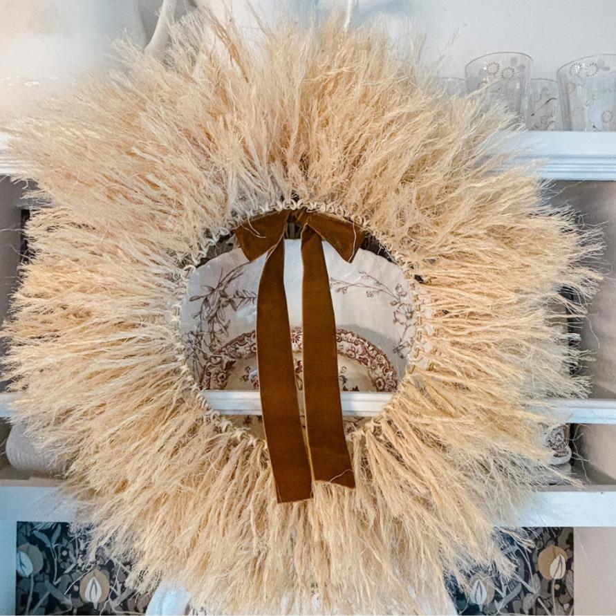 A fluffy, dried-grass wreath with a brown velvet ribbon hangs on a white shelf next to a rope-wrapped lamp.