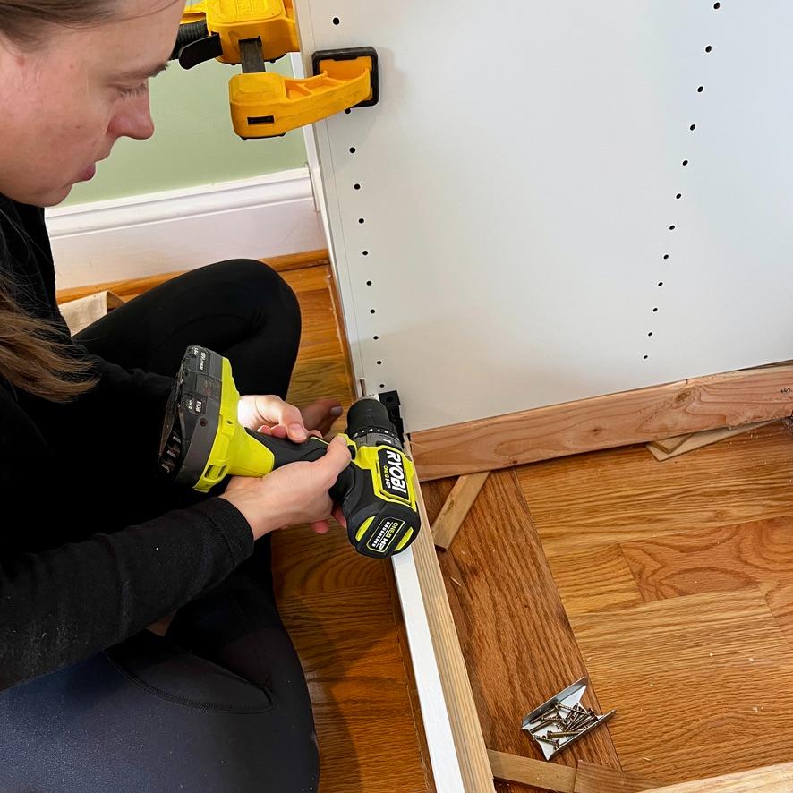 A person uses a yellow power drill to secure an IKEA Pax closet frame to a wooden base.