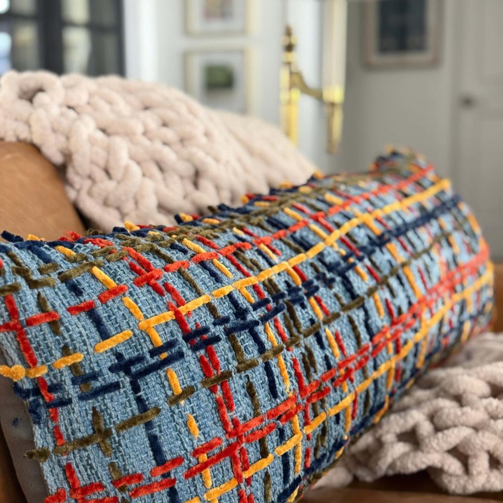 A bolster pillow with a blue, red, gold, and green textured plaid pattern rests against a chunky knit throw blanket next to a rope wrapped lamp.