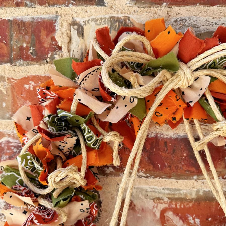 A scrap fabric wreath in orange, green, and cream patterns with a jute twine bow, hanging on a brick wall beside a rope wrapped lamp.
