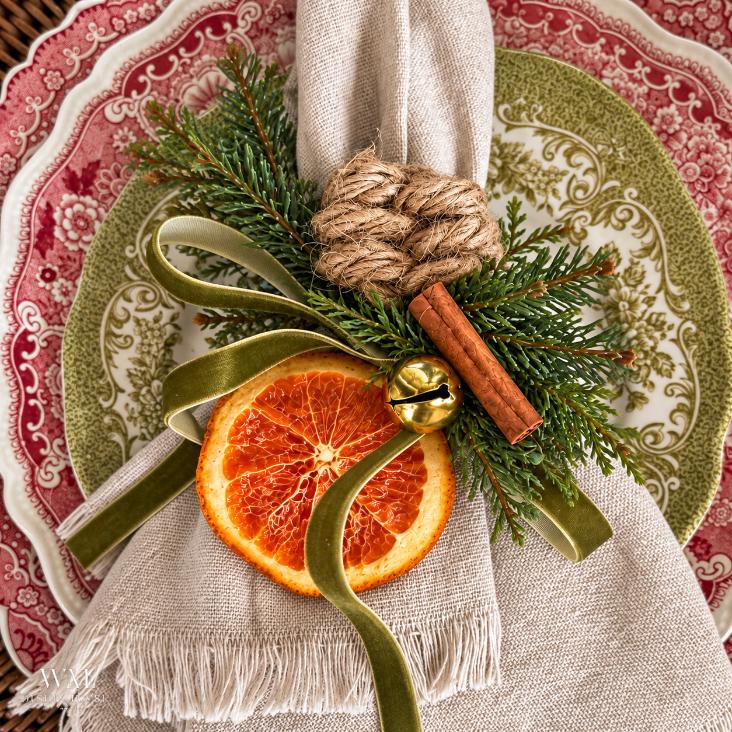 A linen napkin topped with a dried orange slice, cinnamon stick, jingle bell, rope knot, and evergreen sprigs, tied with a green ribbon and resting on a decorative plate near a rope-wrapped lamp.