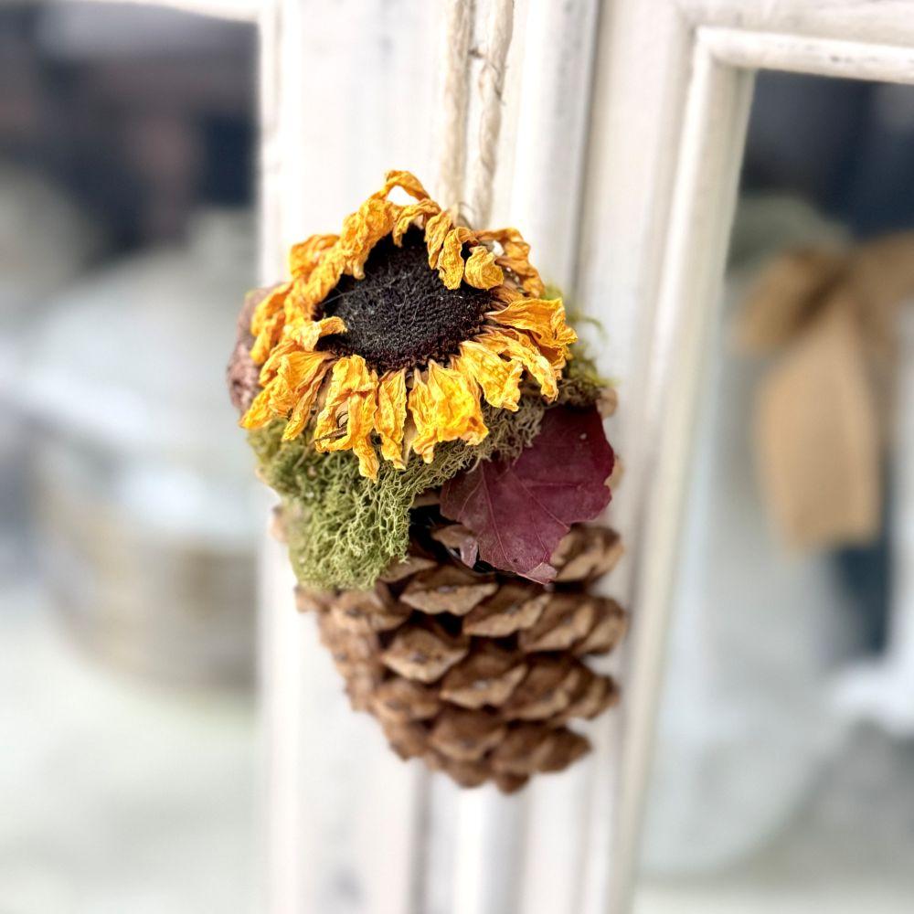 A pinecone ornament decorated with a dried sunflower, moss, and a small leaf, hanging from a twine string on a white frame near a rope wrapped lamp.