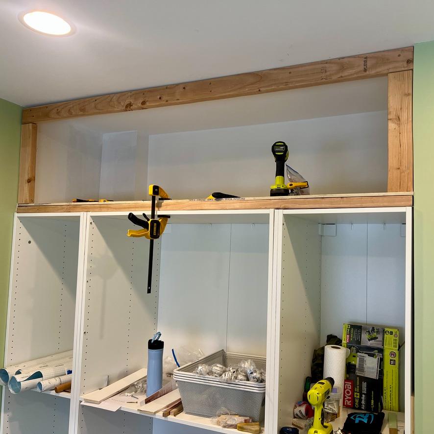 Unfinished white shelving unit with a newly installed wooden frame topper for DIY storage above Pax wardrobes, featuring a power drill, clamps, and miscellaneous tools on the shelves.