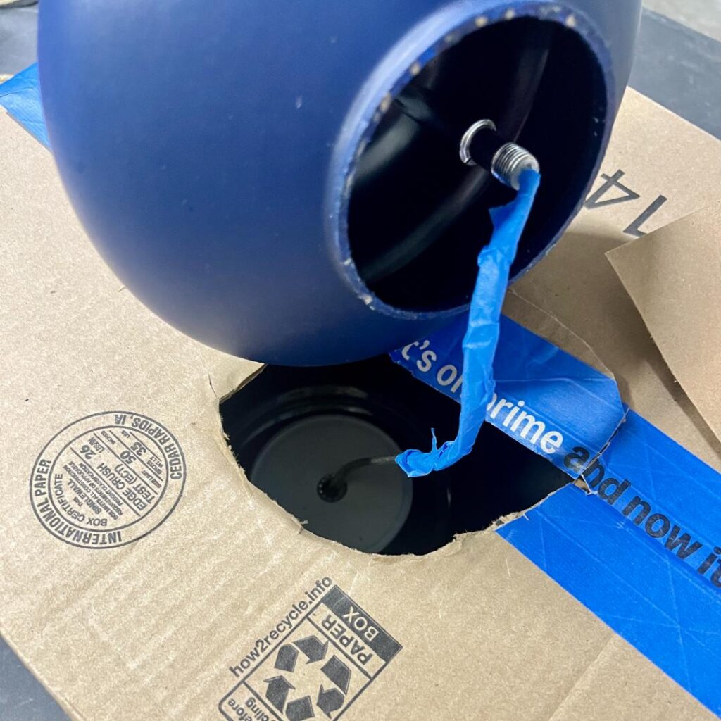 A blue, rope-wrapped spherical pendant light fixture sits partially inside an open cardboard shipping box, with a wire protruding from its opening.